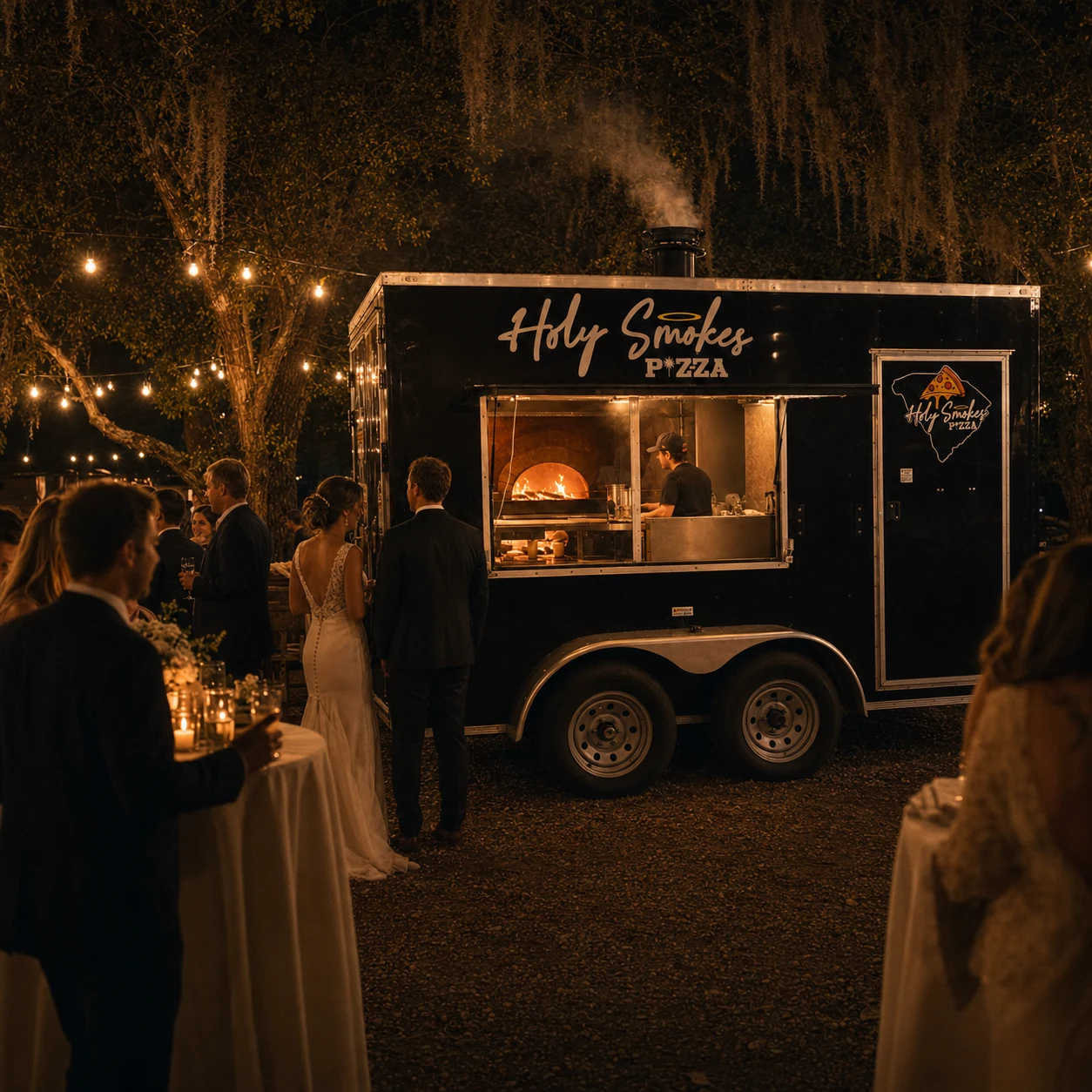 Holy Smokes Pizza serving wood-fired pizza from its branded trailer at an outdoor wedding reception under string lights