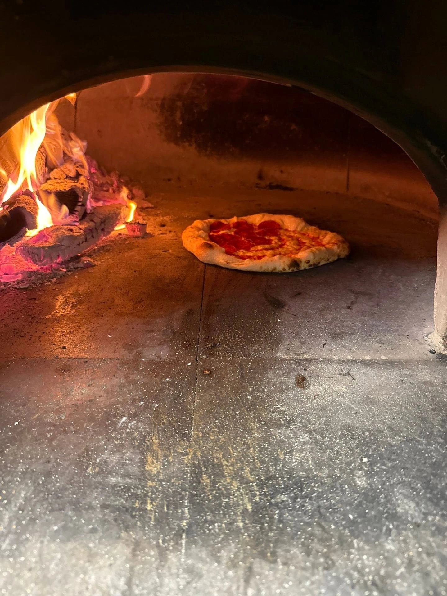 A pepperoni pizza baking beside roaring flames in the wood-fired oven